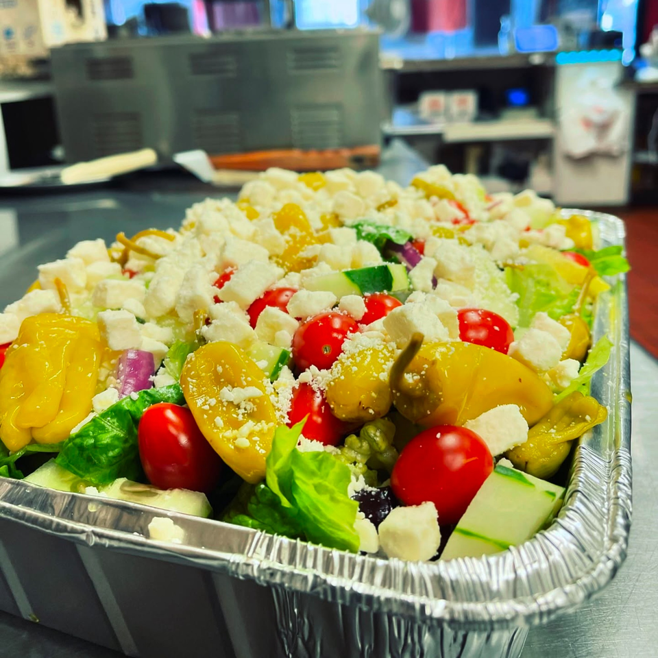 A large Italian salad with lettuce, tomatoes, peppers, cucumbers, and feta cheese in a foil container.