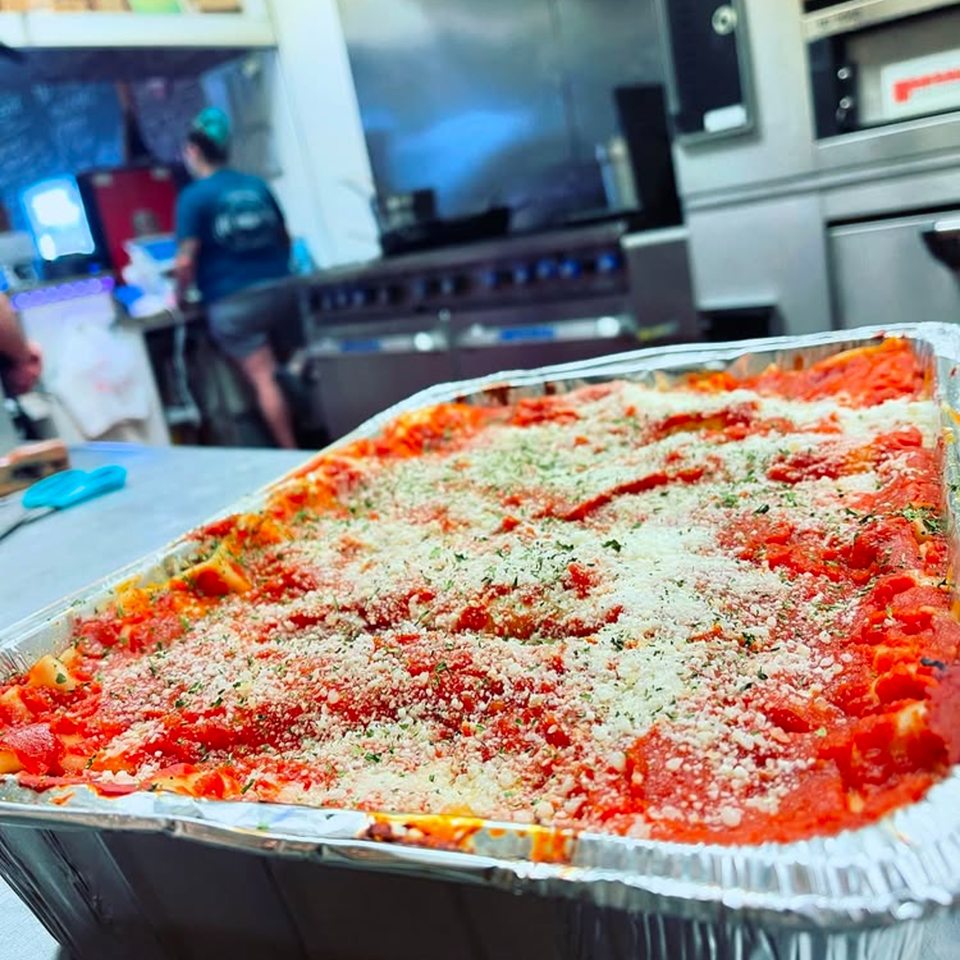A large pan of Anna's Kitchen lasagna, topped with red sauce, parmesan, and herbs, in a kitchen.